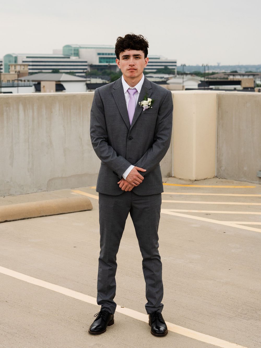 Person in grey suit and purple tie poses formally on rooftop parking area.