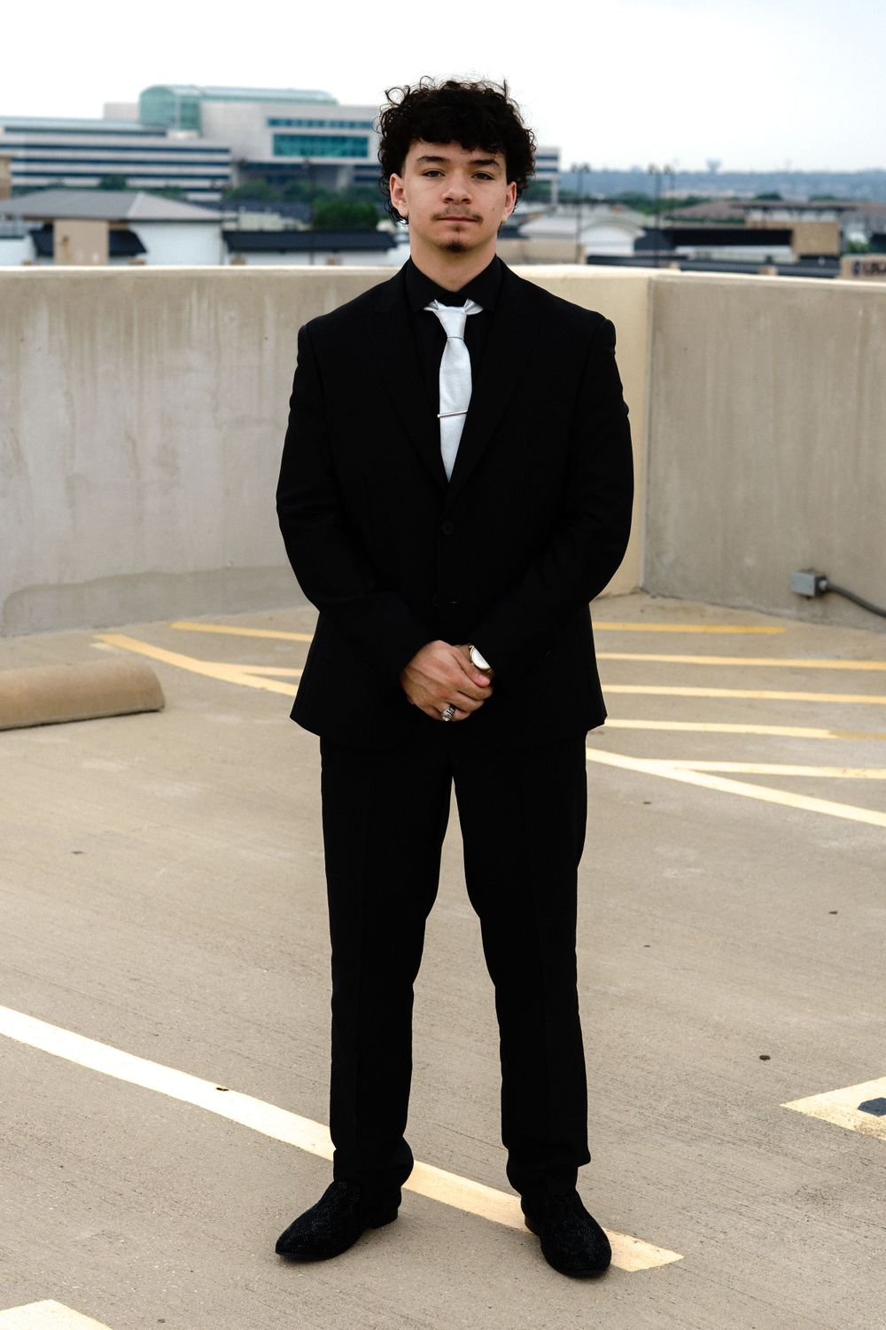 Person in classic black tuxedo poses against rooftop parking lot backdrop.