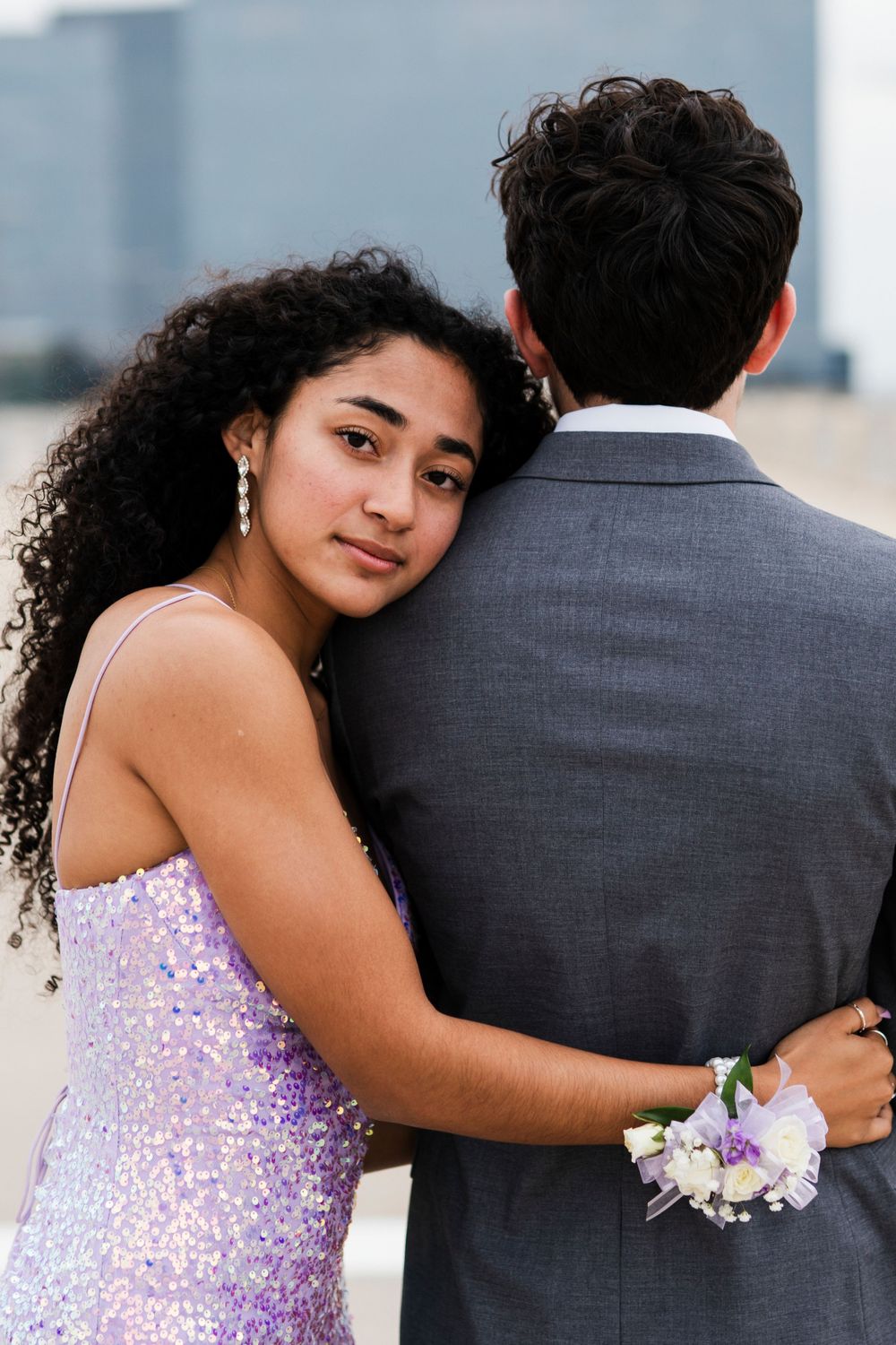 A couple embraces in formal attire with a city skyline backdrop, showing off a purple sequined dress and corsage.