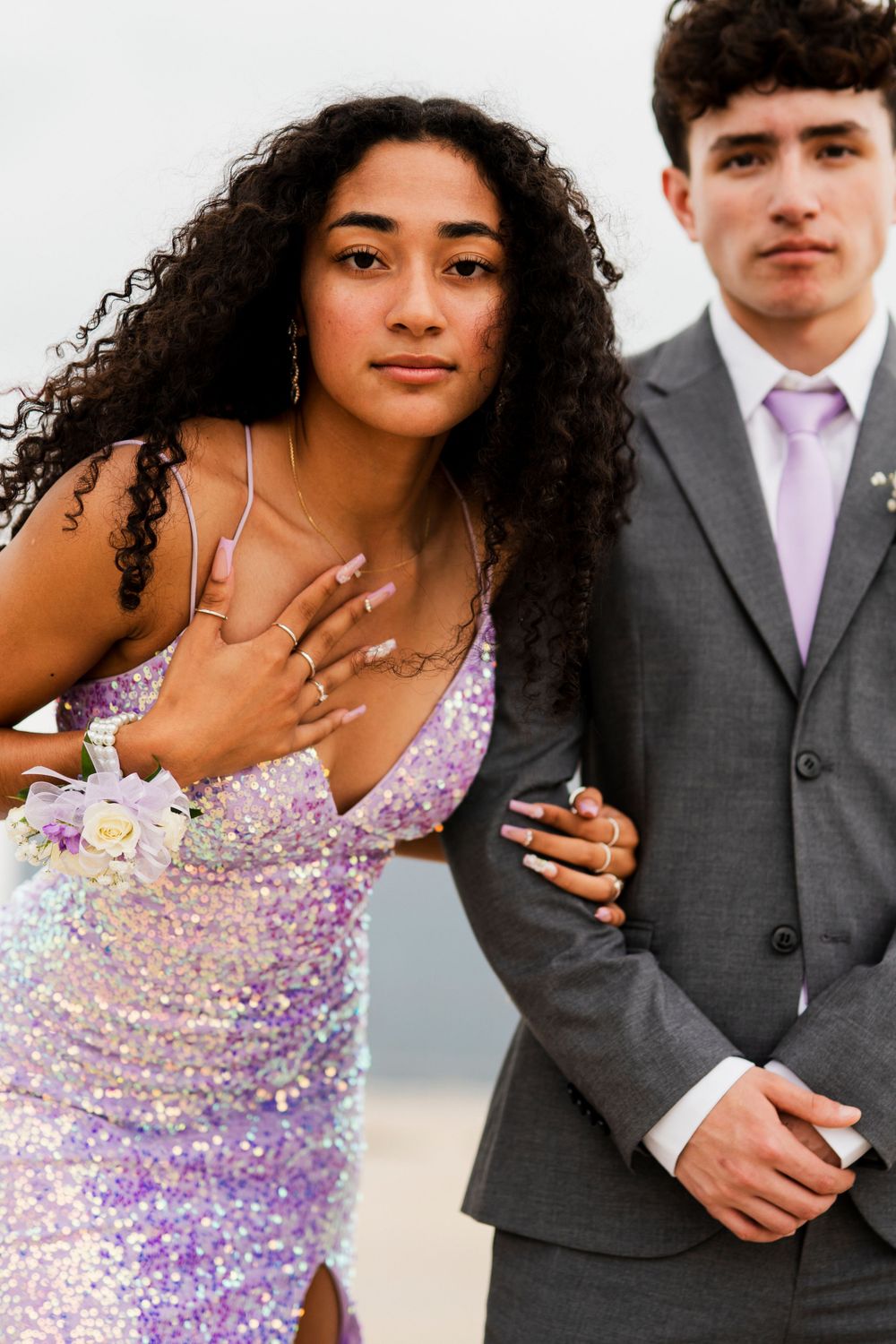 A couple poses in prom attire with the person in purple displaying their corsage.