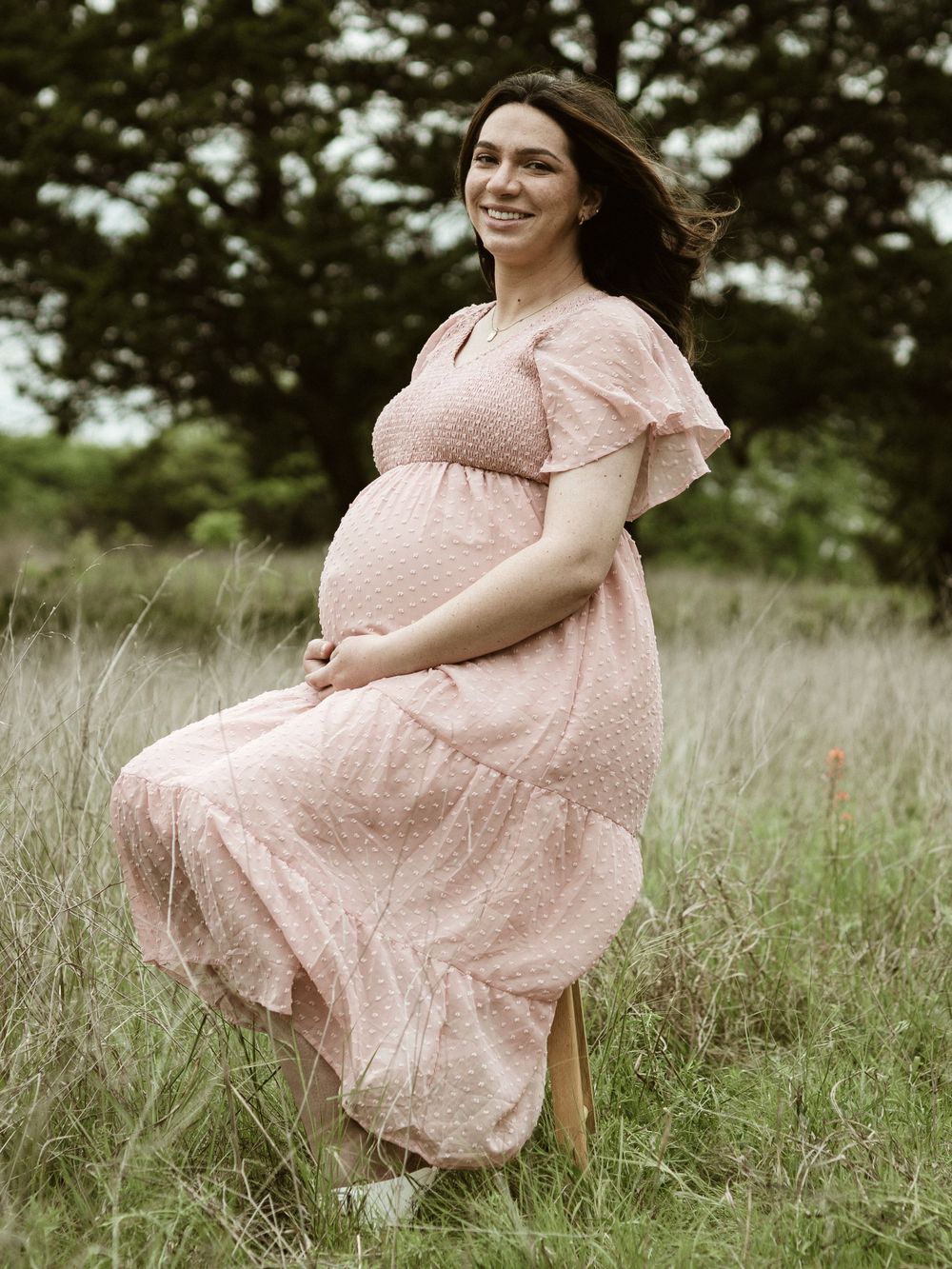 An expectant mother in a pink dress twirls joyfully in a grassy field.