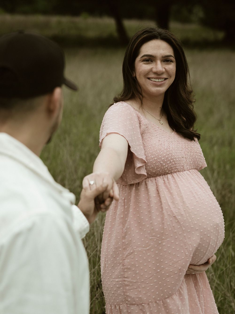Maternity photo in field shows person in pink dress holding hands with partner in white shirt.