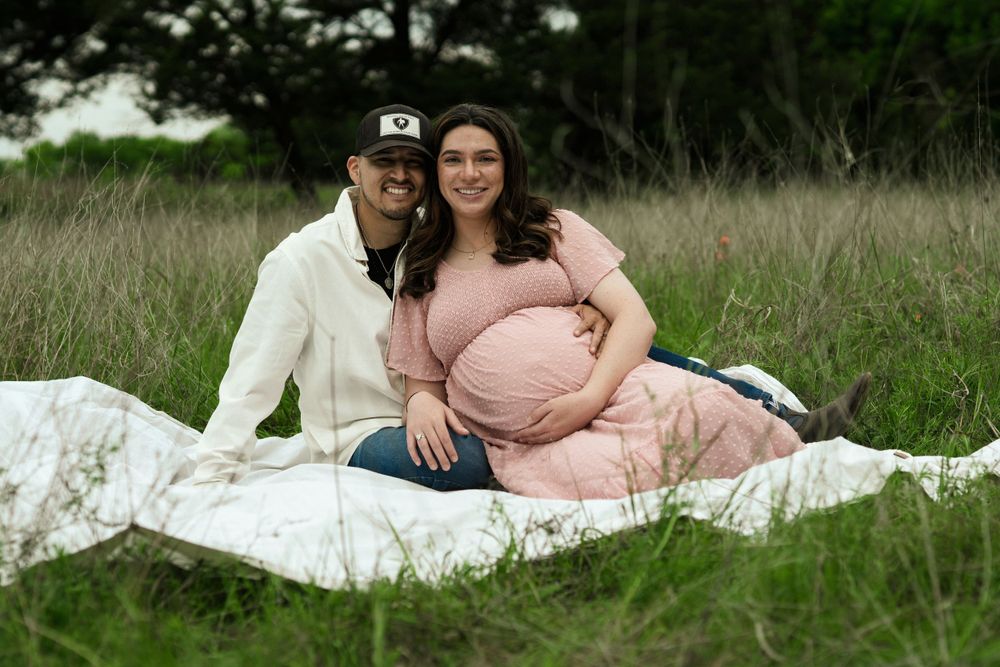 Maternity photoshoot on white blanket in grassy field with couple in coordinated outfits.
