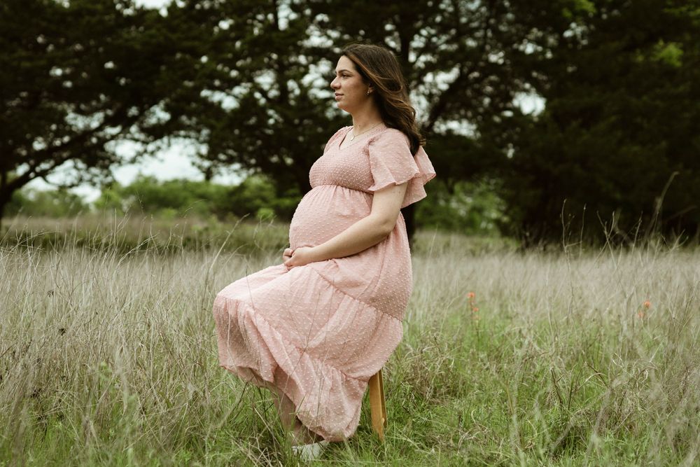Person in flowing pink dress twirls in field during maternity photoshoot.