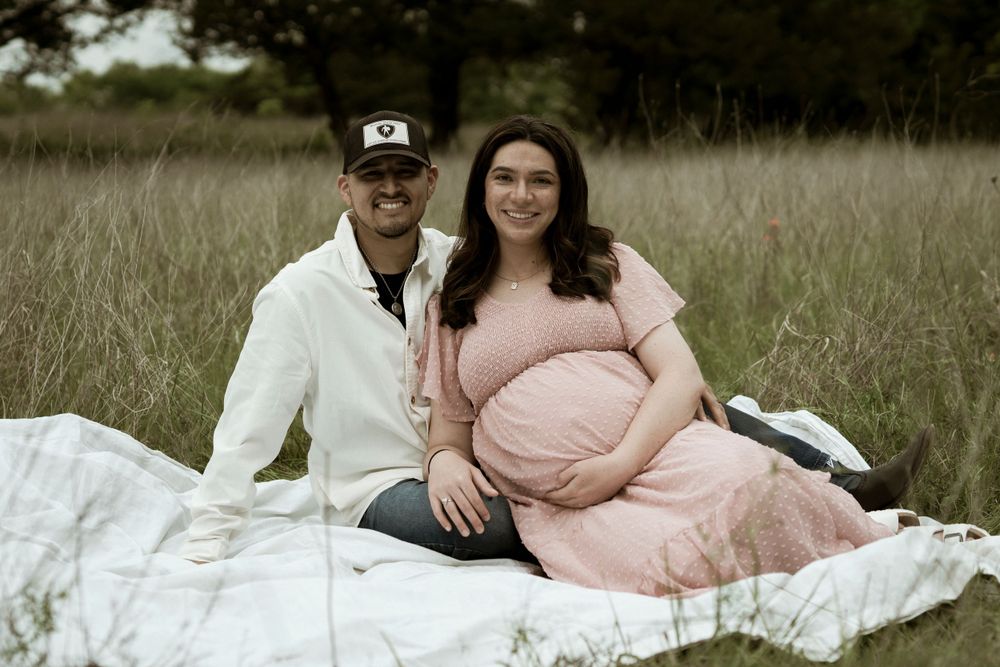 Couple sits on white blanket during outdoor maternity session in grassy field.