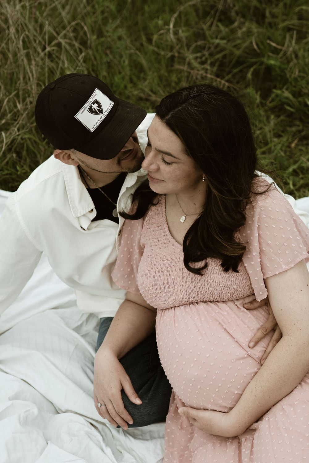 Intimate maternity photo of couple sitting close on white blanket in field.