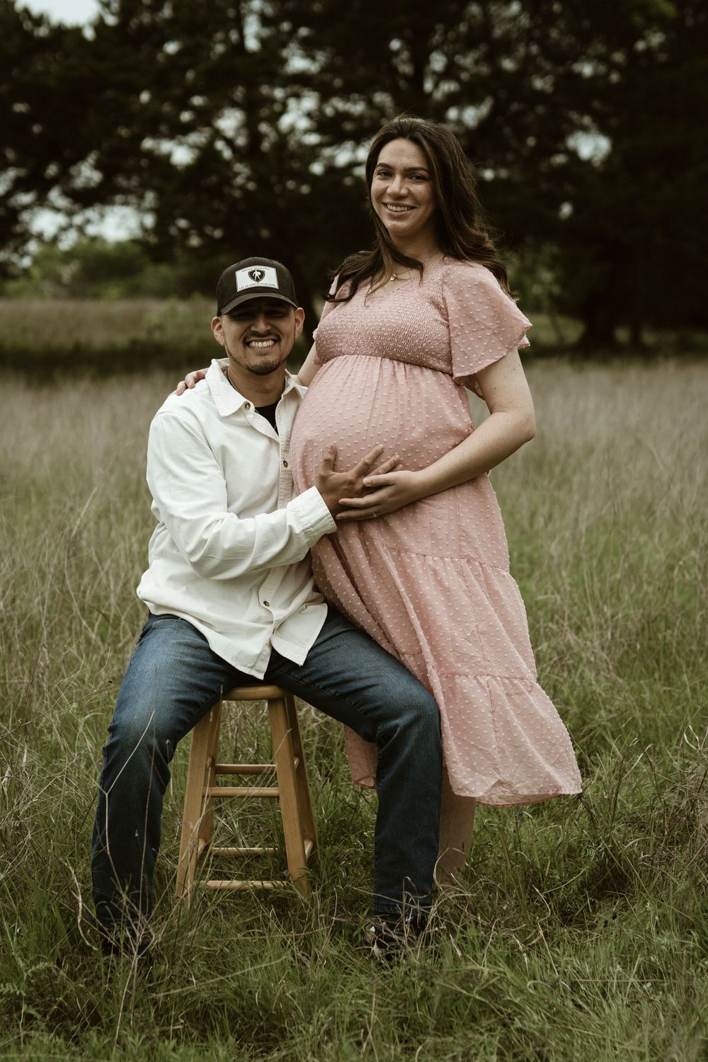 Maternity portrait with person standing beside partner seated on wooden stool in grassy field.