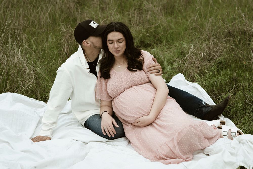 A tender maternity photoshoot in a grassy field with a couple sitting on a white blanket.
