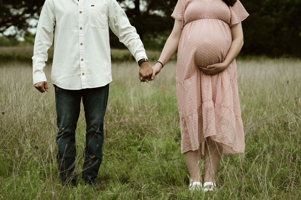 A couple holding hands in a field, with her wearing a flowing pink maternity dress.