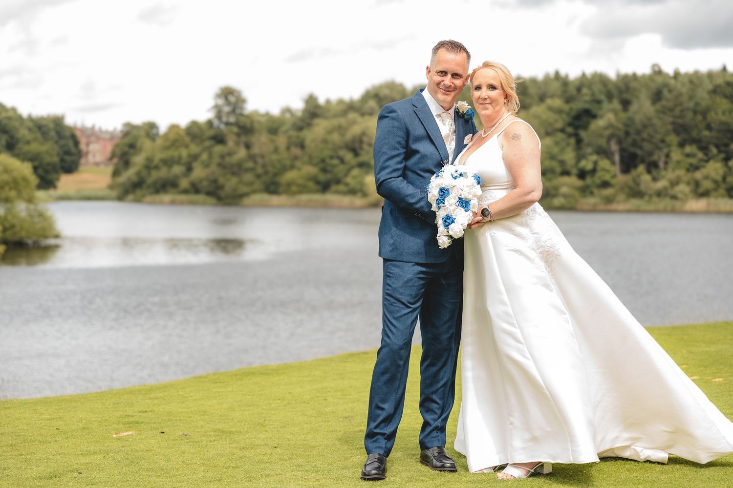 A couple poses with a blue bouquet by a lakeside on their wedding day wearing formal attire.
