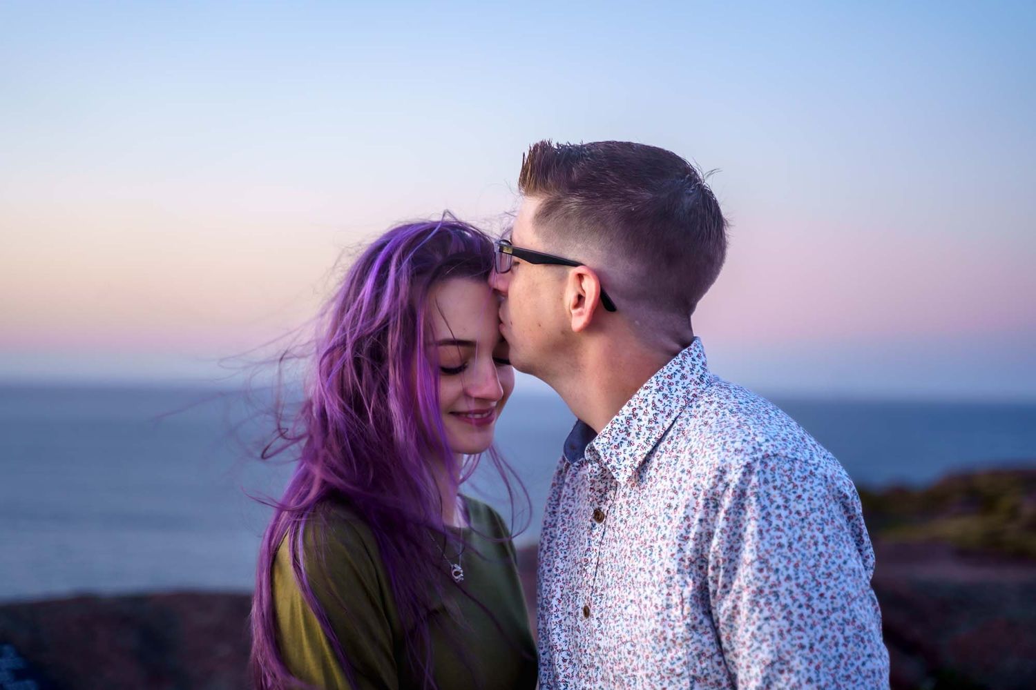 Fiancé kissing fiancée's forehead at sunrise, with a purple sky and ocean view in the background at Cape Spear.