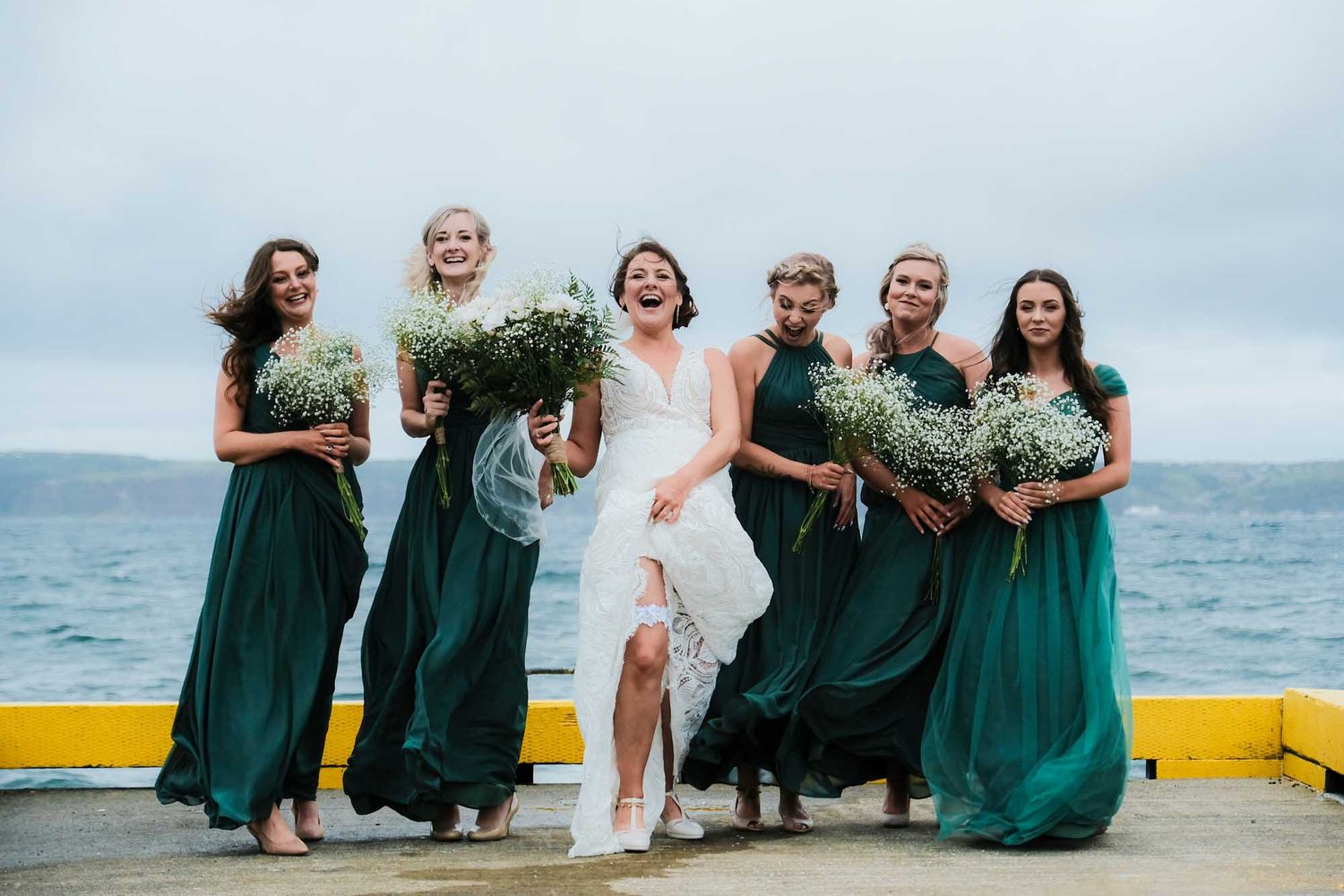 Bride and bridesmaids on a Newfoundland wharf, playfully lifting their dresses to show a hint of leg, St. John's.