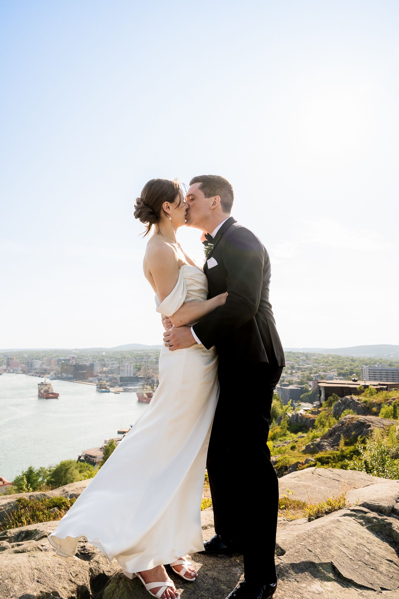 Newlyweds kissing on a cliff above St. John's harbour, soft mist and backlit hazy sky behind them, Newfoundland.