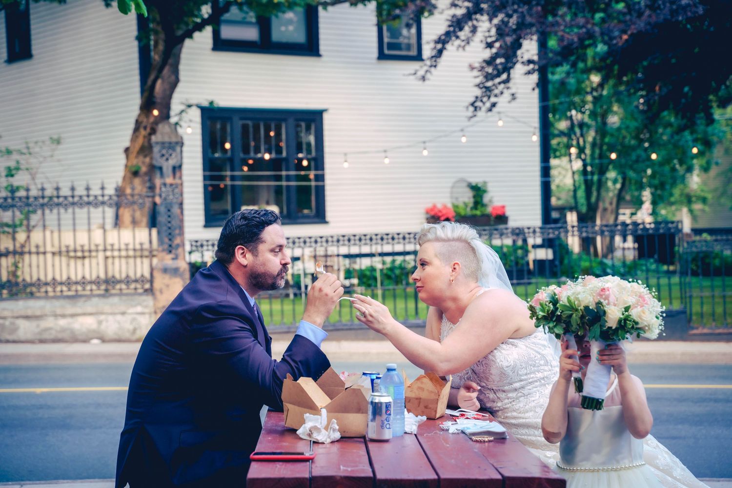 Newlyweds sharing poutine on the street after their wedding ceremony, a small child peeking through flowers nearby, St. John's, Newfoundland