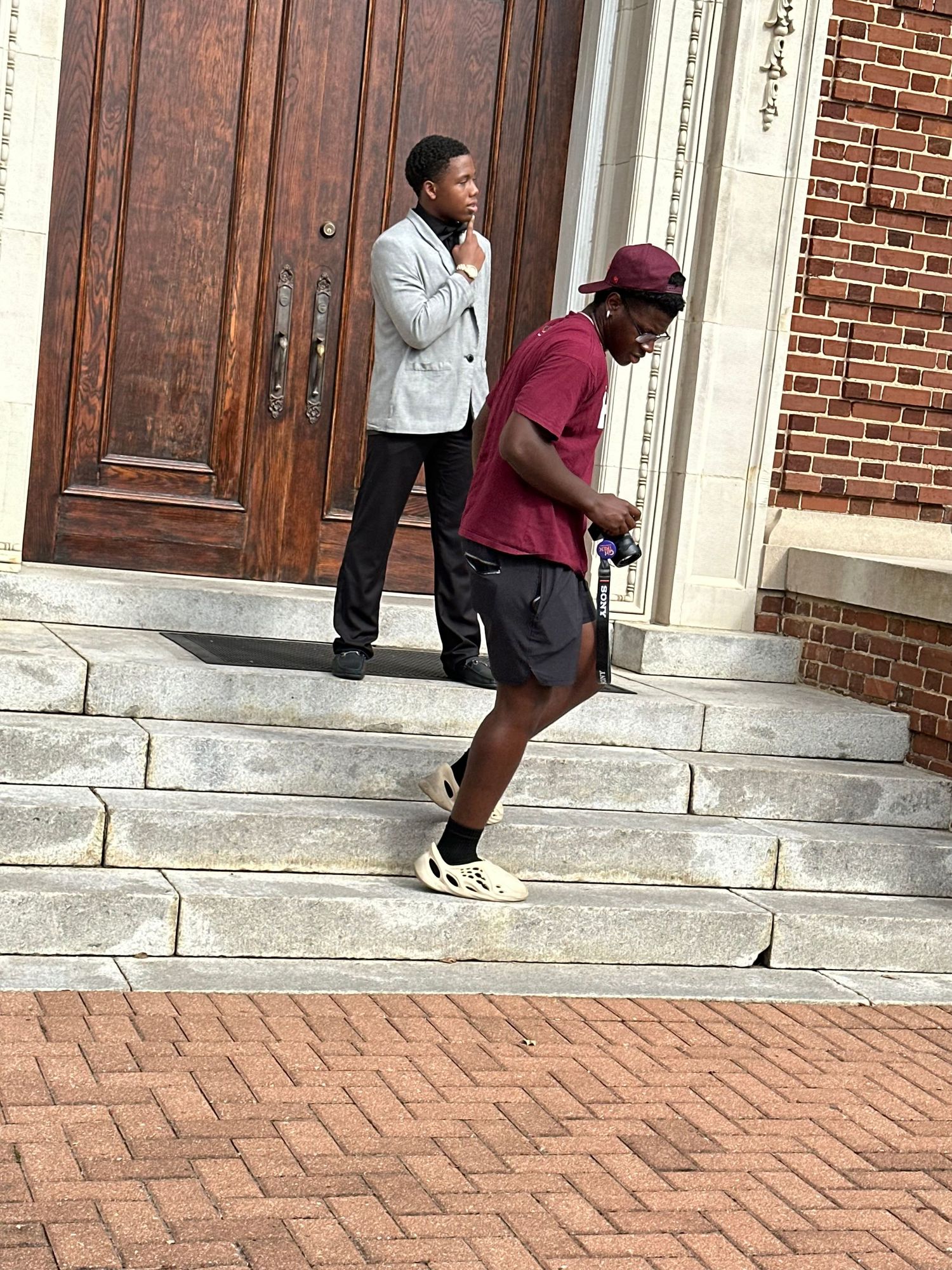 Two young men pose outdoors on stone steps in front of a large wooden door, one in a gray blazer and one in a maroon shirt holding a camera.