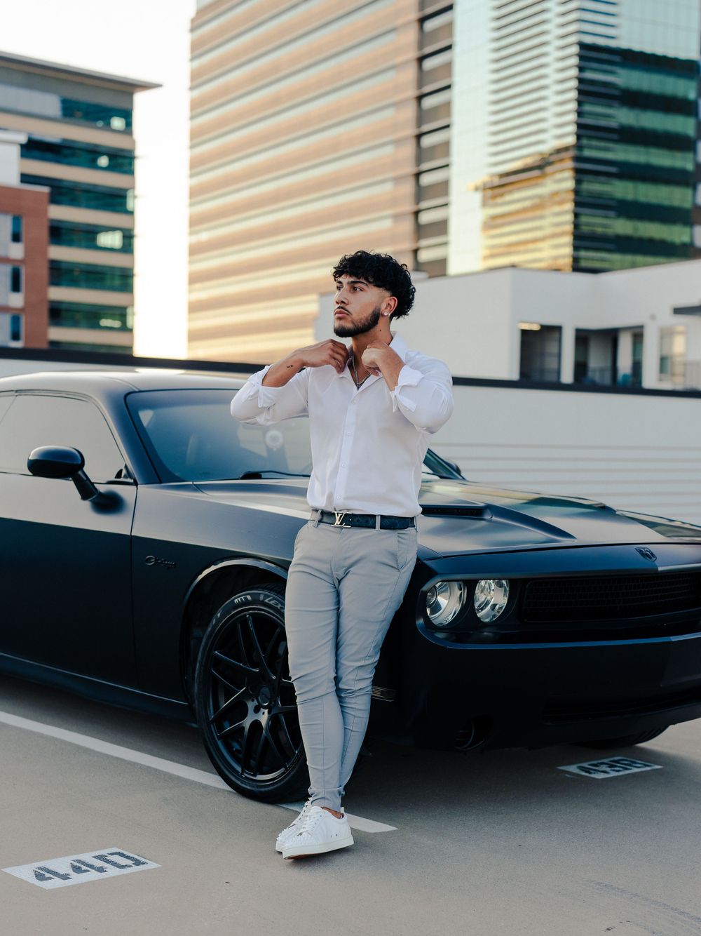 Stylish man in white shirt and grey pants poses beside black muscle car in urban setting.