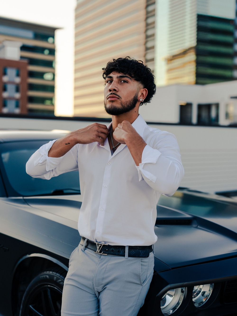 Fashionable outfit with white button-down shirt and grey pants posed against black Dodge Challenger in urban setting.
