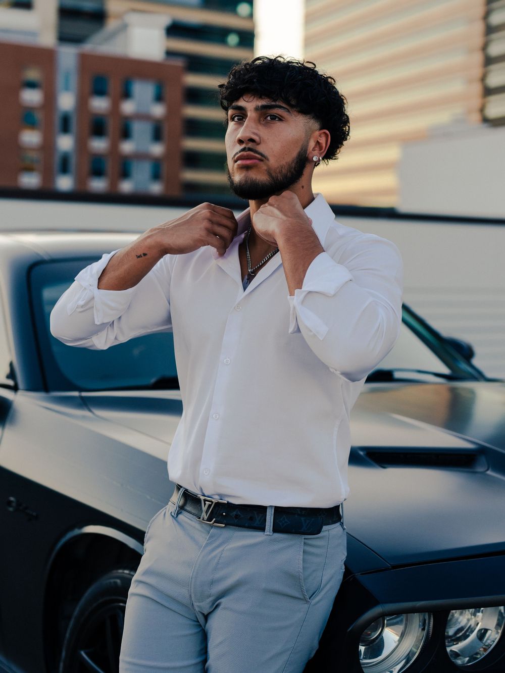 Fashion portrait of man adjusting collar against black car and city skyline.