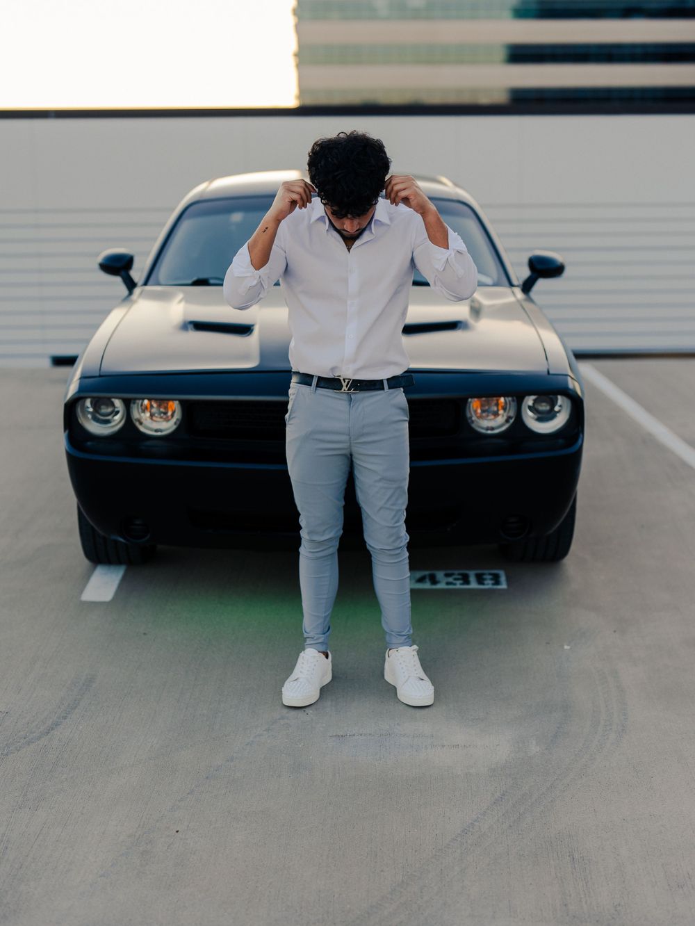 Urban fashion pose featuring monochrome outfit against black muscle car in parking garage.