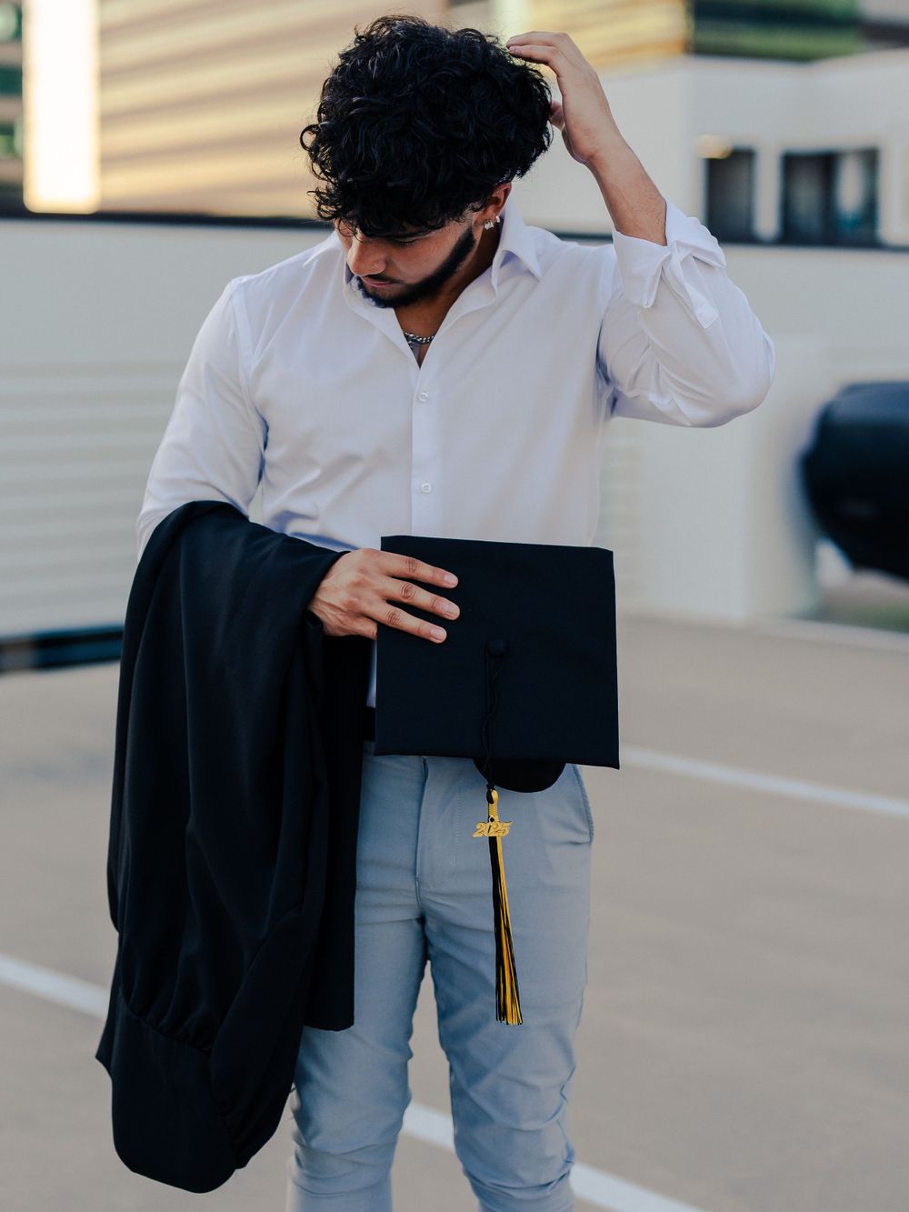 Graduate in formal attire holds graduation cap and gown in parking structure.