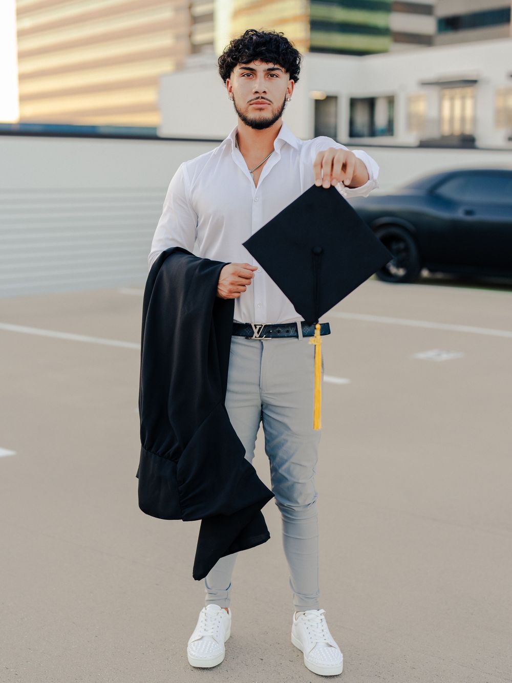 Graduate poses with graduation cap and black gown against urban backdrop.
