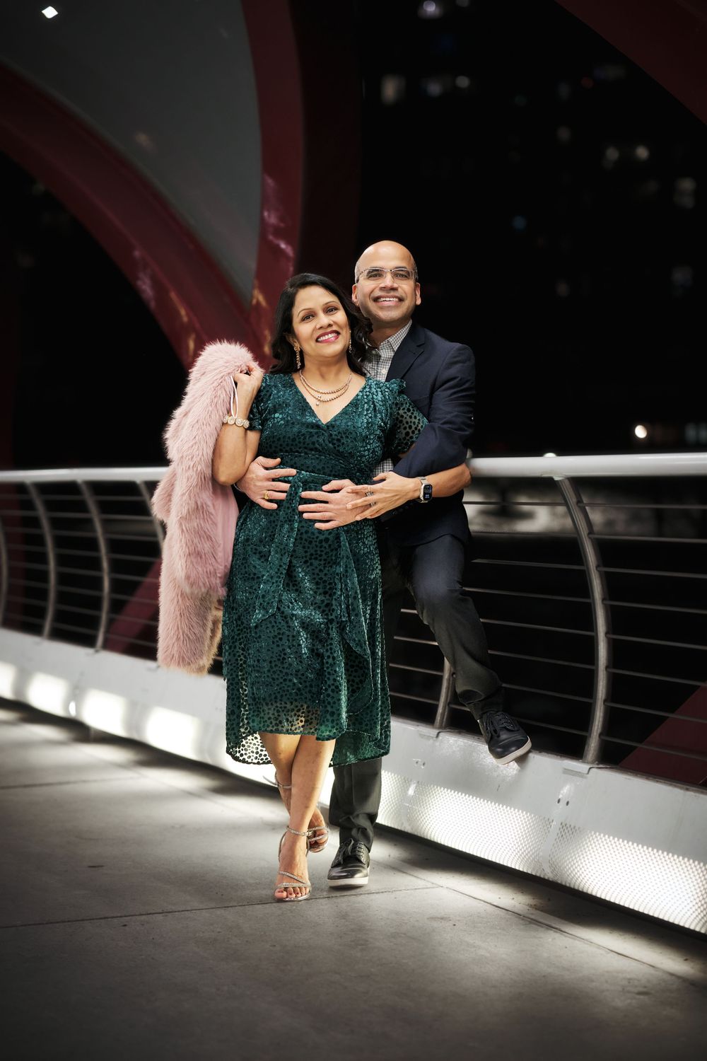 A couple poses on a walkway at night, with the woman wearing a sparkly green dress and pink coat.