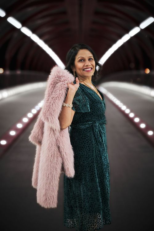 A person in an emerald green dress and pink fur coat poses in an illuminated tunnel with curved walls and lighting.