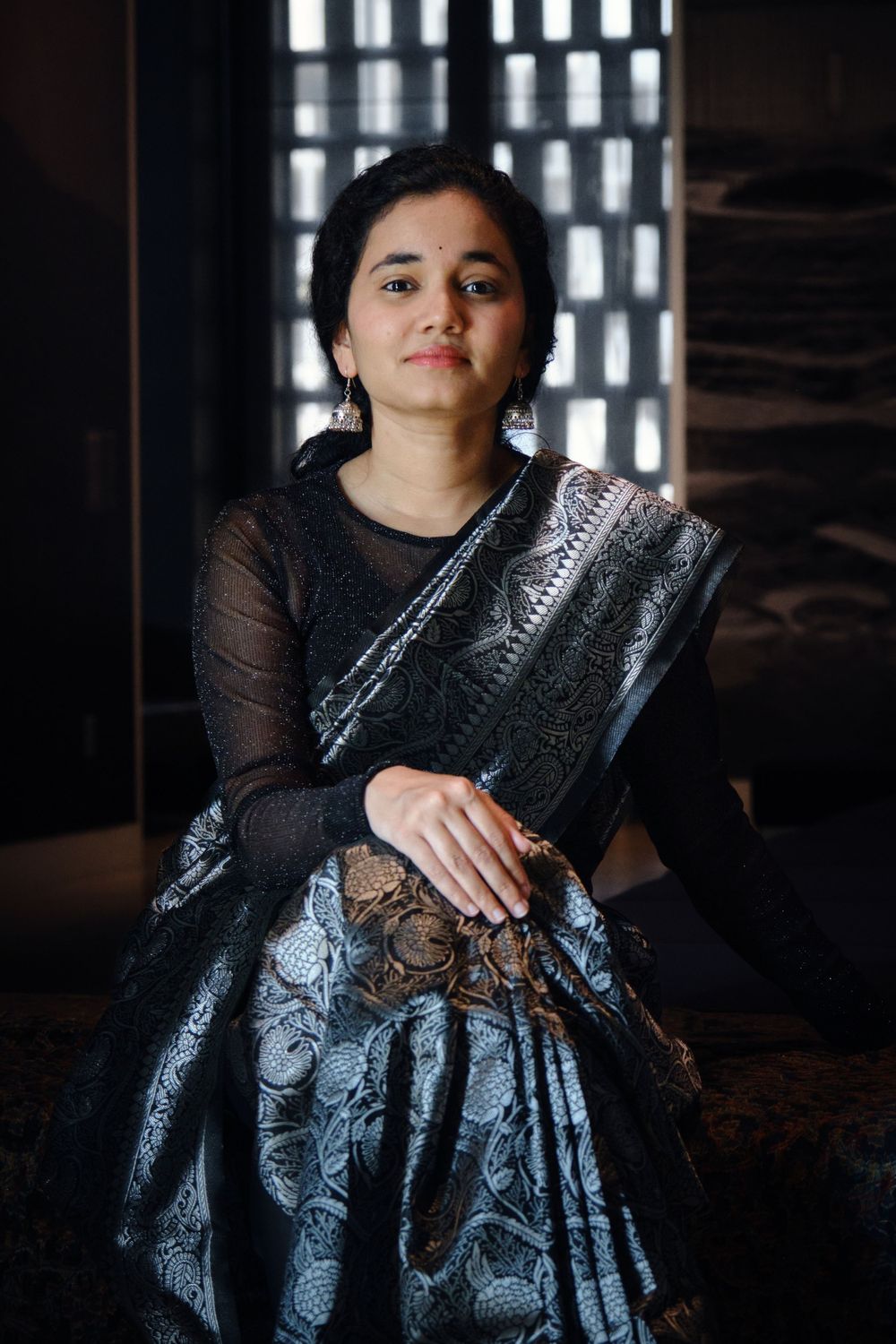 Portrait of person in black and silver sari seated against geometric window backdrop.