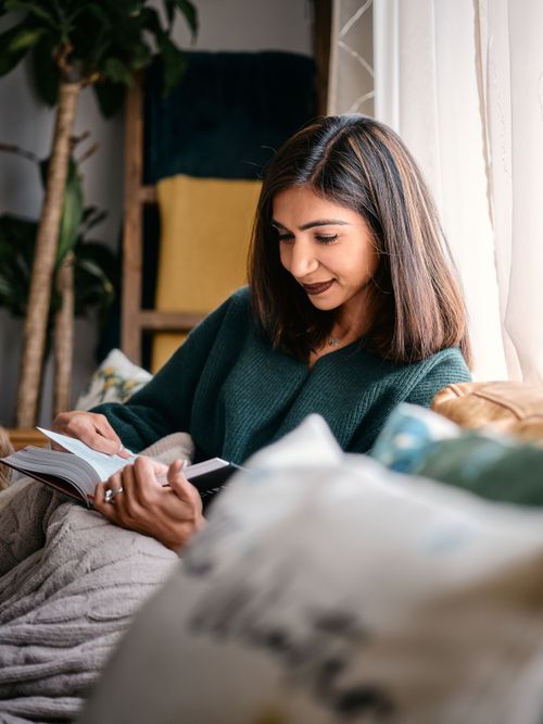 Someone relaxes on a couch reading a book by natural window light while wearing a cozy dark green sweater.