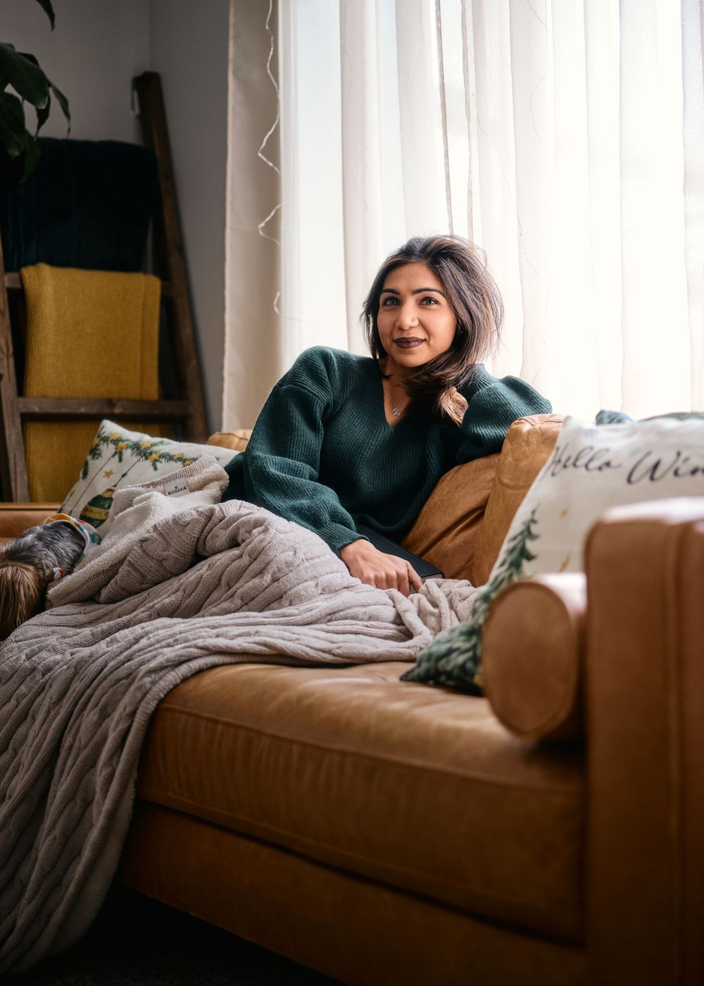 Person relaxing on tan couch with blanket wearing green sweater in cozy living room setting.