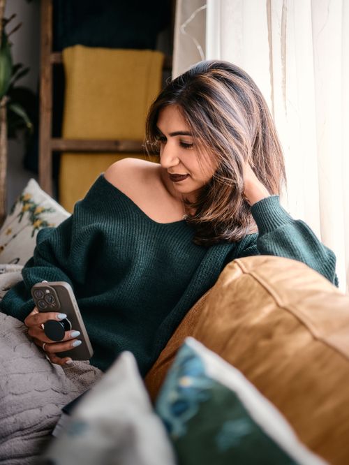 Person in emerald green sweater relaxing on couch with phone and cushions by window in cozy setting.