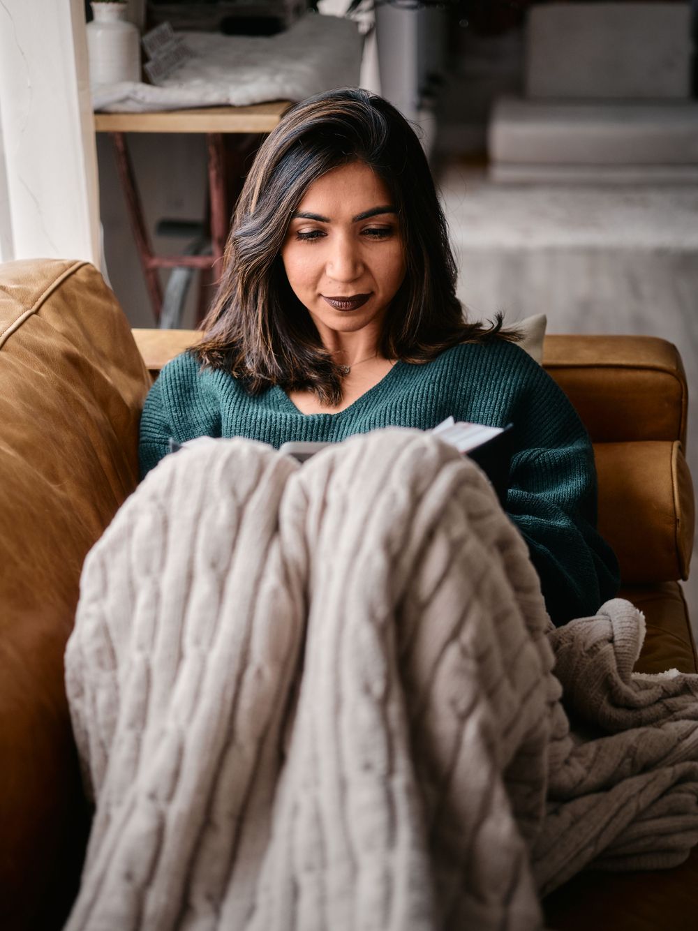 Someone relaxing on a leather couch wearing a green sweater while wrapped in a cozy cable knit blanket.