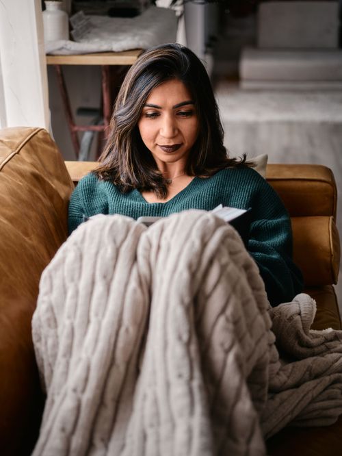 Someone relaxing on a leather couch wearing a green sweater while wrapped in a cozy cable knit blanket.