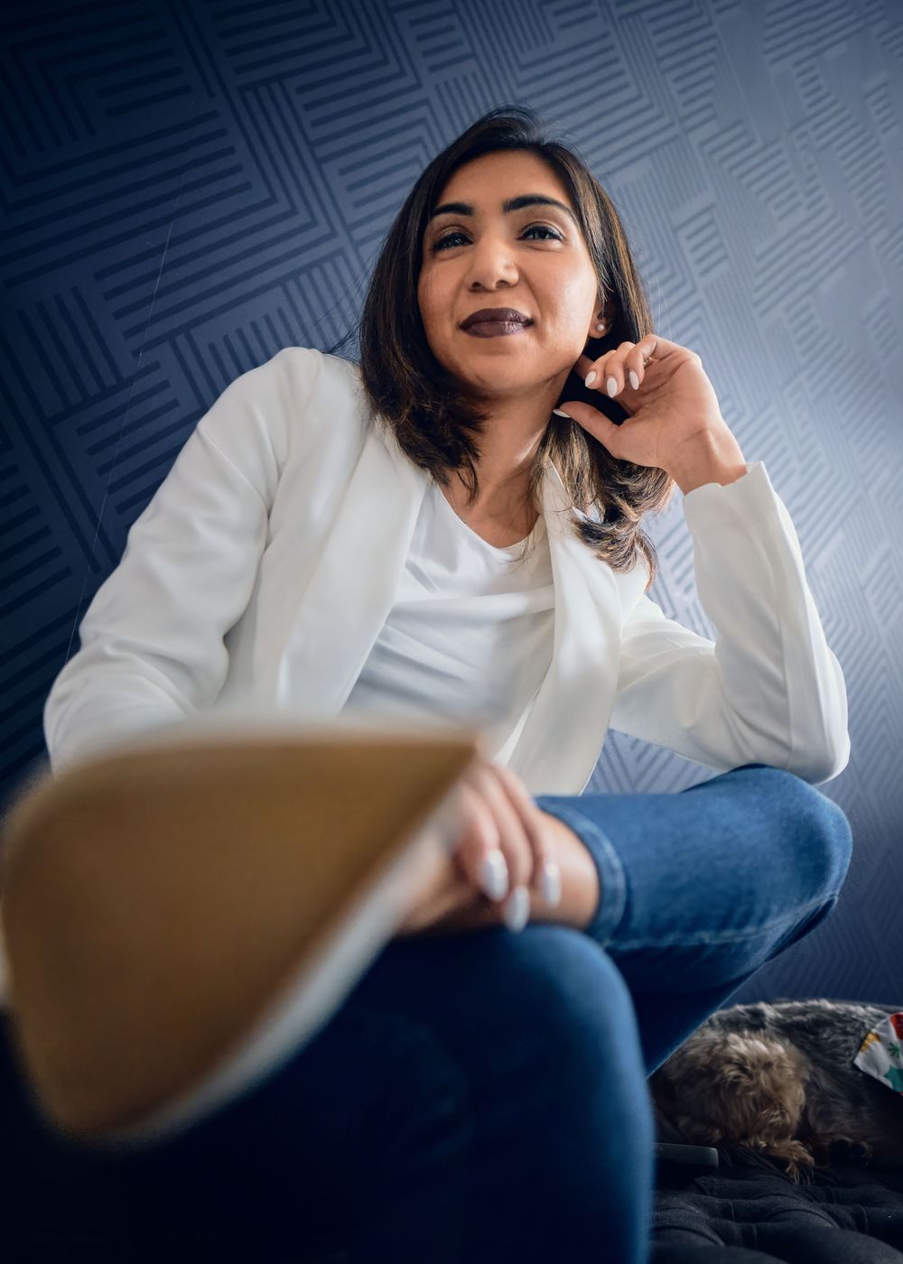 A person wearing a white blazer and jeans sits casually on a couch against a blue patterned wall.