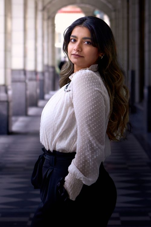 A woman in a white sweater and dark skirt poses elegantly against a backdrop of architectural columns.