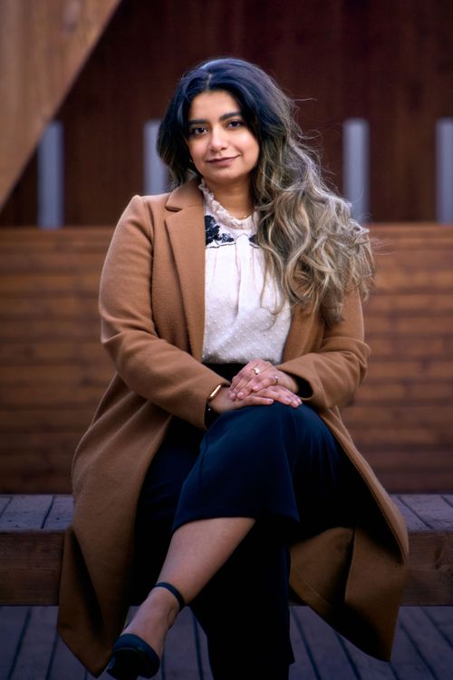 A person in a brown coat and dark skirt sits on wooden steps, with long wavy hair styled in an ombre effect.