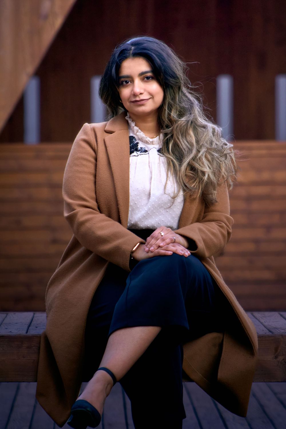 A person in a brown coat and dark skirt sits on wooden steps, with long wavy hair styled in an ombre effect.