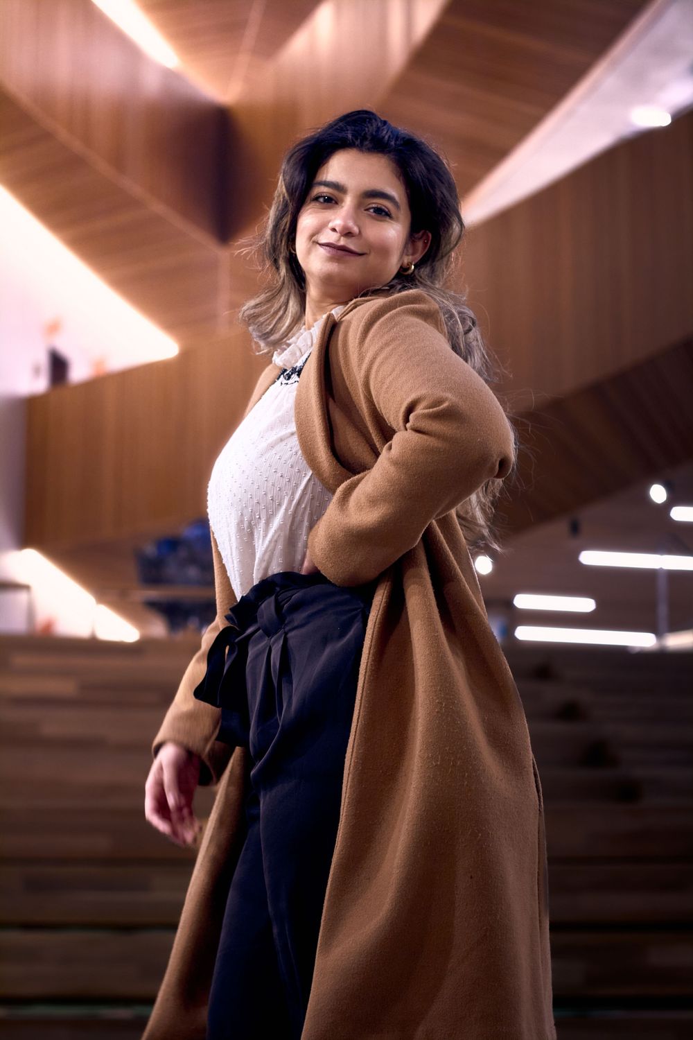 A person poses confidently in a tan cardigan, white top and navy pants under dramatic lighting in an urban setting.