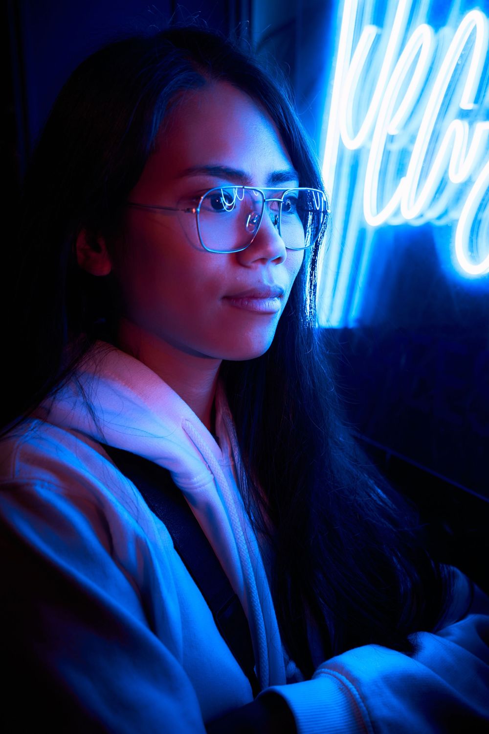 Portrait in neon lighting showing someone wearing glasses and a white hoodie with blue illumination reflecting off their face.