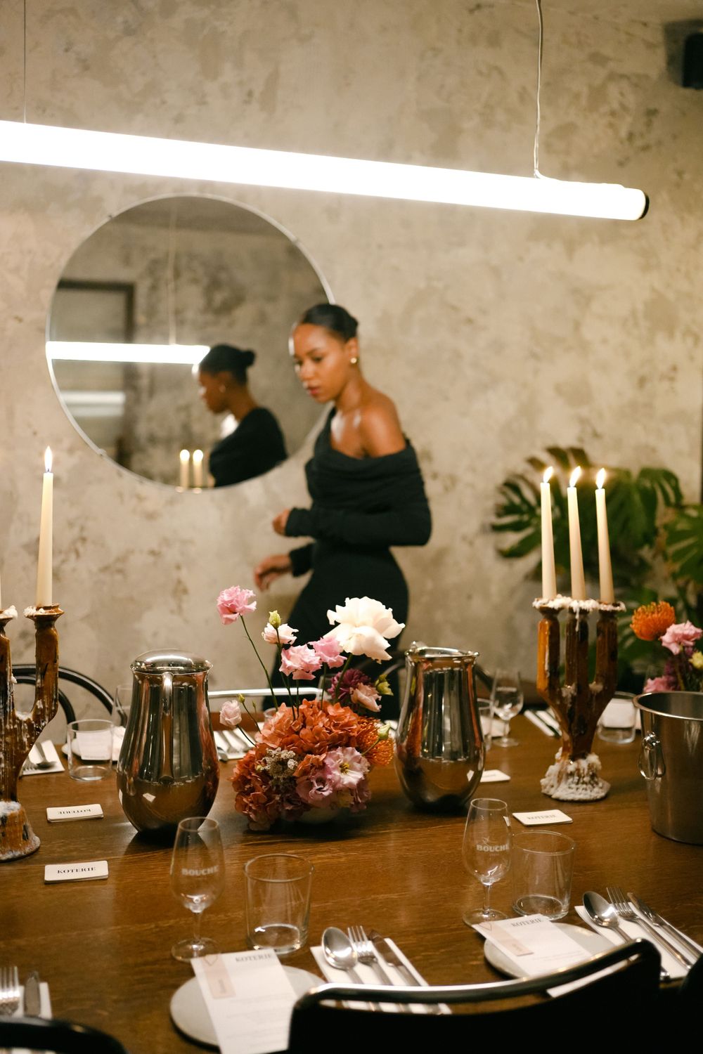 Modern restaurant table setting with floral arrangements, candles and round mirrors on textured walls.