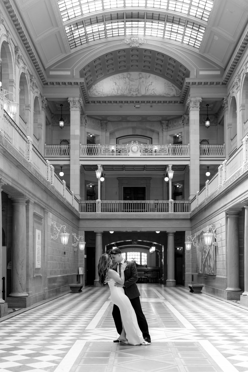 An elegant couple dancing in a spacious neoclassical hall with vaulted ceilings and white marble floors.