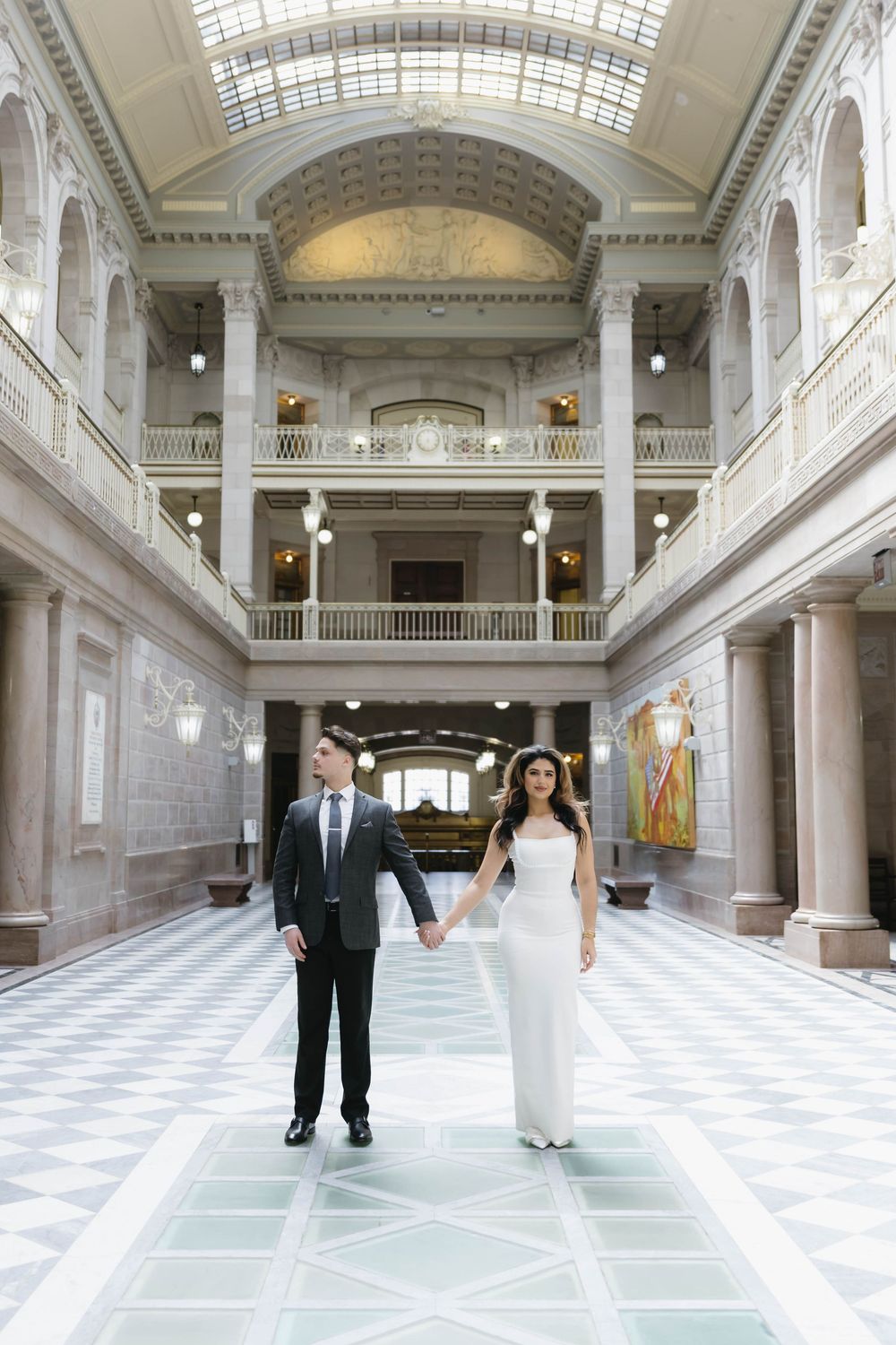 A couple shares an intimate moment in a majestic hall with classical columns and natural skylight.