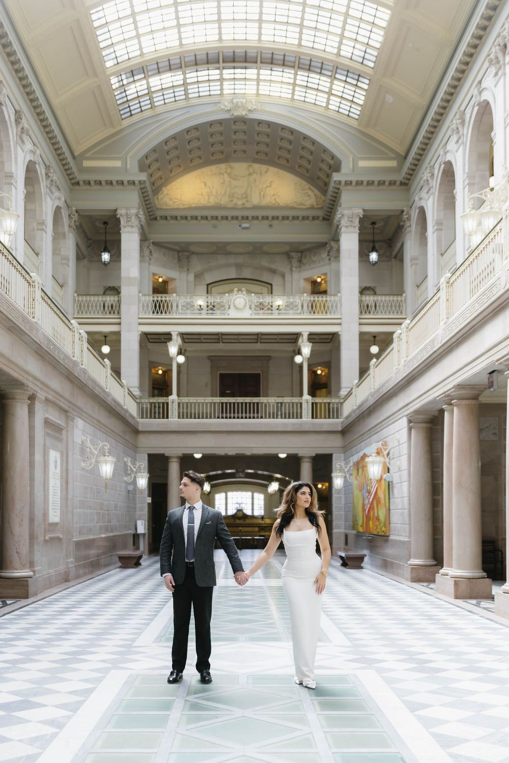 A couple poses in a grand hall featuring ornate architecture and dramatic natural lighting.