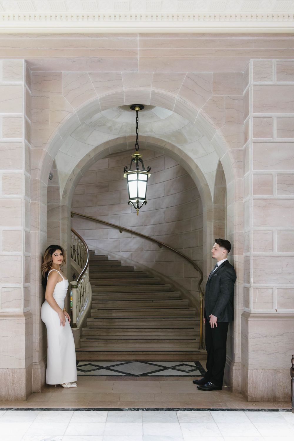 A couple stands in an elegant stone archway with dramatic staircase and pendant lighting.