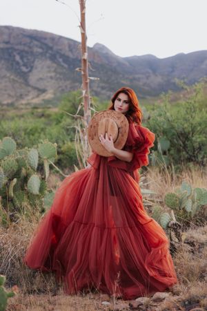 Voluminous burgundy tulle dress contrasts with desert cacti and mountain scenery in artistic portrait.