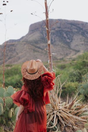 Vintage-inspired fashion shoot features flowing red dress and wide-brimmed hat against mountain landscape.