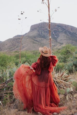 Desert fashion portrait showcases flowing red dress and hat against mountainous backdrop.