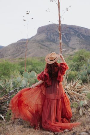 Ethereal red dress billows in desert landscape while model poses with vintage hat.