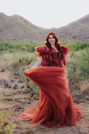 A voluminous burgundy gown swirls elegantly in a desert landscape with mountains.