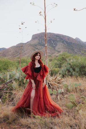 Fashion portrait captures flowing red dress and hat among desert brush with mountain vista.